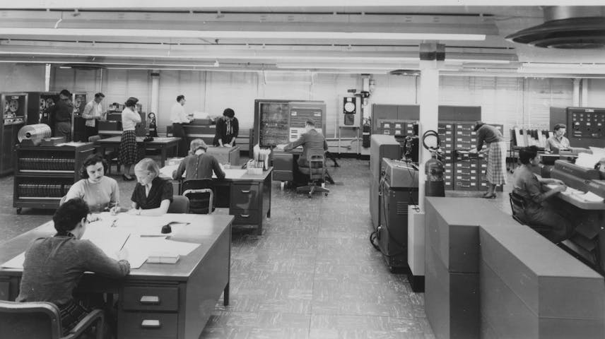 Historical photograph of several women seated at desks with papers, calculators, and office equipment in a workroom.