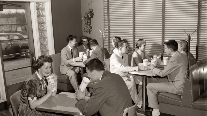A black and white photo from the 1950s showing a group of teenagers hanging out at a diner or soda shop. They are sitting at small tables and in booths with shiny vinyl seats, drinking beverages through straws from paper cups. The girls are wearing dresses and the boys are in pants and jackets. A 1950s car is visible through the window in the background.
