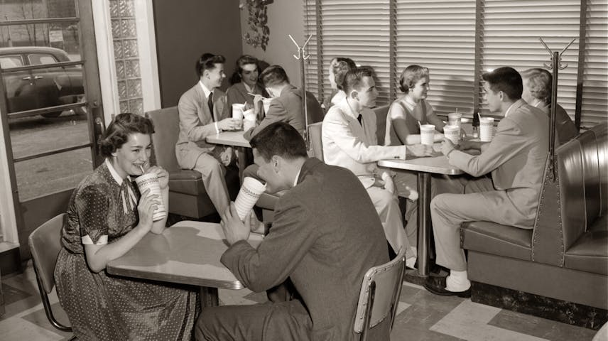 A black and white photo from the 1950s showing a group of teenagers hanging out at a diner or soda shop. They are sitting at small tables and in booths with shiny vinyl seats, drinking beverages through straws from paper cups. The girls are wearing dresses and the boys are in pants and jackets. A 1950s car is visible through the window in the background.