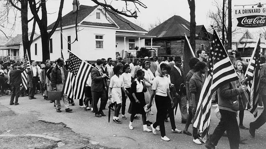A black-and-white photograph showing a large group of people marching down a residential street, carrying American flags. The marchers, who are Black and white men, women, and children dressed in everyday clothing, walk together in a peaceful protest. In the background are modest houses and a Coca-Cola sign.