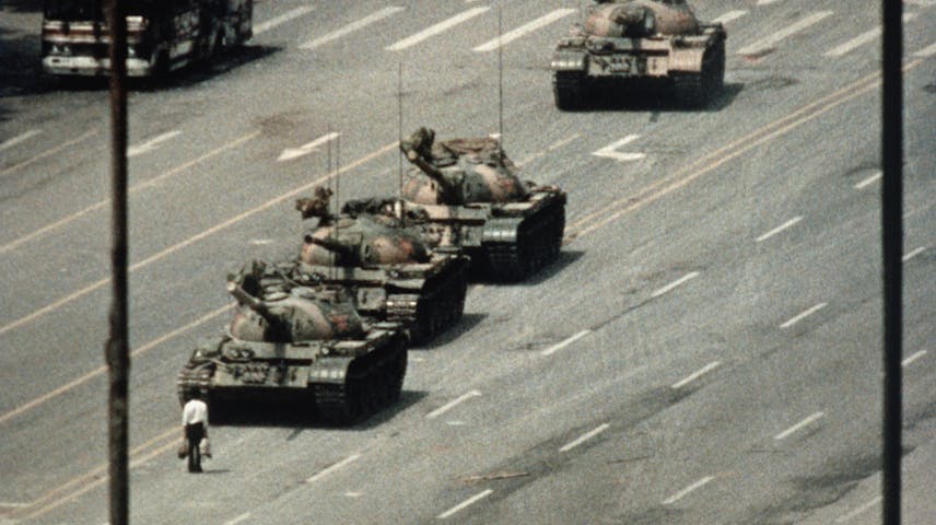 A man stands in the middle of a street blocking the path of a column of tanks going down the street.