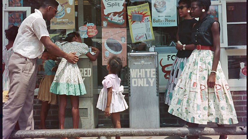 A color photograph from the 1950s showing a group of Black Americans — a man, two young girls, and two teenage girls — standing outside a Dairy Queen ice cream shop. In the center of the image is a drinking fountain labeled WHITE ONLY, a sign used during racial segregation in the American South to prevent Black people from using the same facilities as white people. The storefront displays cheerful ice cream advertisements, creating a painful contrast with the discriminatory sign. This photograph documents the everyday reality of Jim Crow laws, which enforced racial separation in public spaces.
