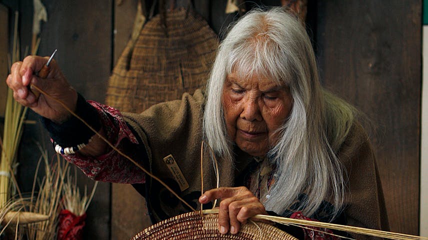 Elderly Native American woman sits weaving a basket by hand.