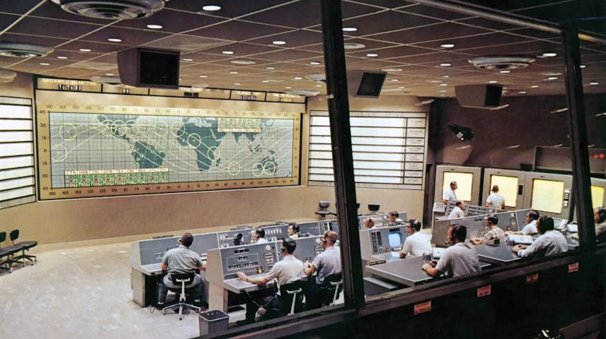 Photograph of a Flight Control room of NASA. Through large windows and from behind can be seen rows of computer stations manned by personnel wearing headphones. On the large wall in front of them is a flat map of the world.