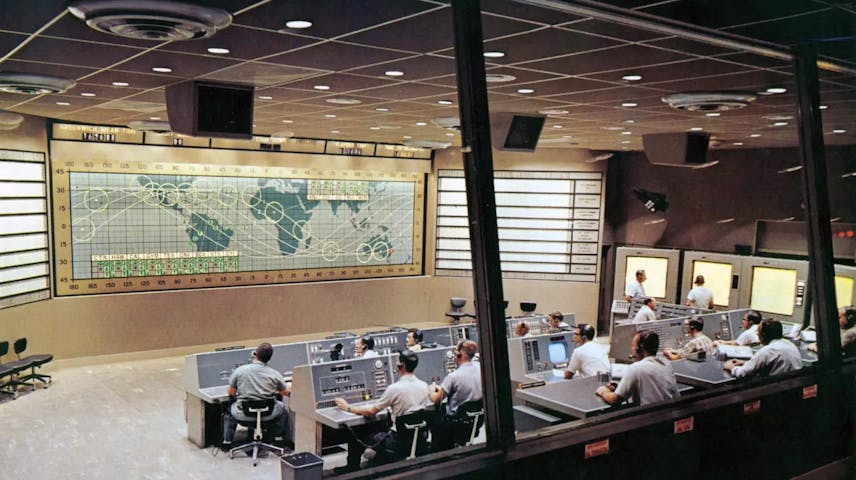 Photograph of a Flight Control room of NASA. Through large windows and from behind can be seen rows of computer stations manned by personnel wearing headphones. On the large wall in front of them is a flat map of the world.