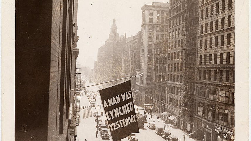 A black and white photo showing a black flag hanging from the window of a tall building in New York City in 1938. The words on the flag say “A man was lynched yesterday” in all capital letters.