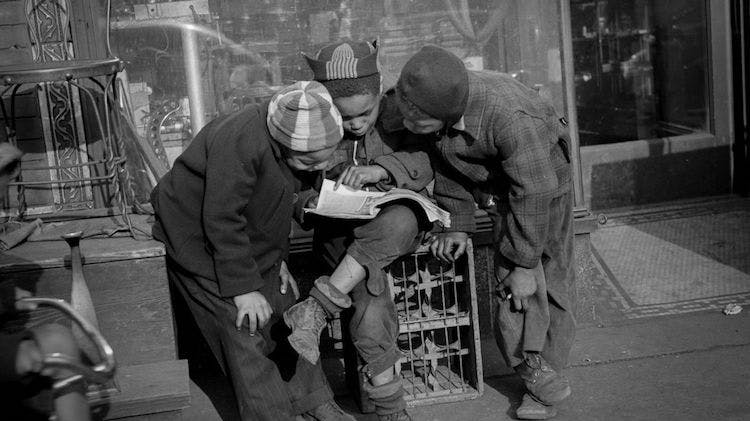 A black and white photo taken in 1941 of three Black boys outside a shop on a city street. The boy in the middle sits on a crate while the other two peer over his shoulder at the magazine he is reading.