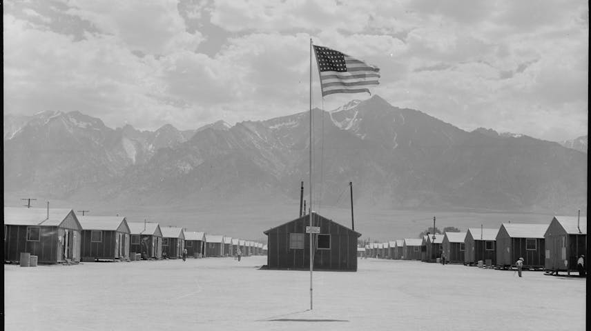 A black-and-white photograph showing rows of plain, identical wooden barracks stretching into the distance on both sides of a flat, dusty open space. An American flag flies from a tall flagpole in the center of the image. In the background, a rugged mountain range with snow on its peaks rises against a cloudy sky. A few small figures of people can be seen walking between the buildings. This is a photograph of a Japanese American incarceration camp during World War II, where tens of thousands of Japanese Americans were forced to live after the U.S. government removed them from their homes following the attack on Pearl Harbor in 1941.