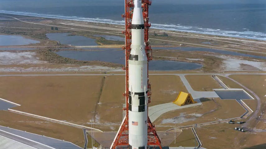 Photograph of a massive rocket being held upright by a red tower structure sitting atop a launch platform. The rocket is mostly white and is decorated with the American flag and “USA.” In the background are the beach and ocean.