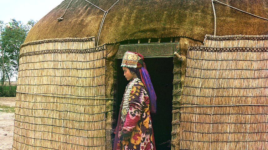 Full-length profile portrait of an Uzbek woman, dressed in traditional clothing and jewelry, standing on a richly decorated carpet at the entrance to a yurt.