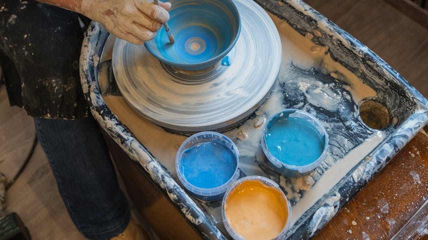 Image of a potter at a pottery wheel painting blue glaze onto a bowl.
