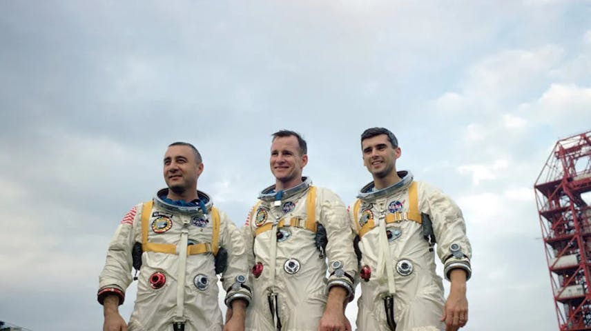 Three men (Virgil “Gus” Grissom, Edward White II and Roger Chaffee) in white astronaut gear pose for a photo with a NASA vehicle in the background.