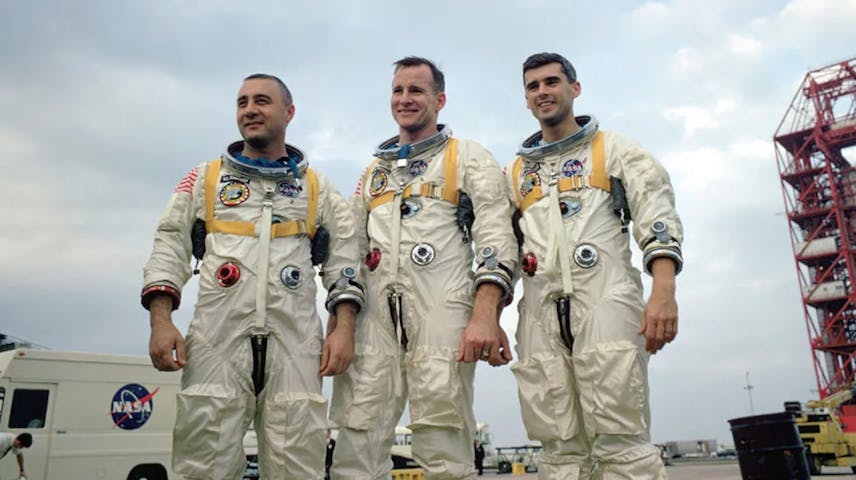 Three men (Virgil “Gus” Grissom, Edward White II and Roger Chaffee) in white astronaut gear pose for a photo with a NASA vehicle in the background.
