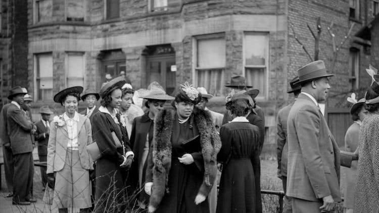 A black and white photo taken in 1941 showing over a dozen Black men and women on a city sidewalk in front of a large brick building. The men wear suits and hats. The women wear dresses, heels and hats, and one wears a fur stole. Most are smiling.