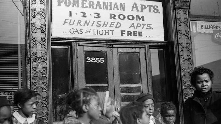 A black and white photo taken in 1941 showing several Black children and one adult woman standing outside an apartment building with a sign advertising “Pomeranian Apartments 1-2-3 Room Furnished Apts. Gas and Light Free”
