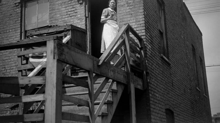 A black and white photo taken in 1941 of a Black woman wearing a daydress and a long white apron. She stands at the top of a wooden staircase in the second story doorway of a brick building.