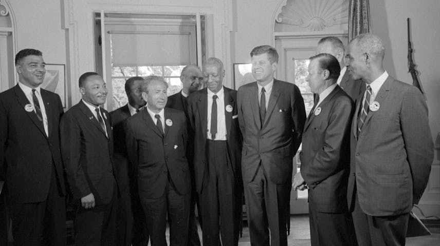 A black-and-white photograph from the 1960s showing a group of men in suits gathered together indoors, smiling and talking. Several of the men are wearing small round buttons or badges on their lapels. It is in the white house with tall windows and decorative door frames.