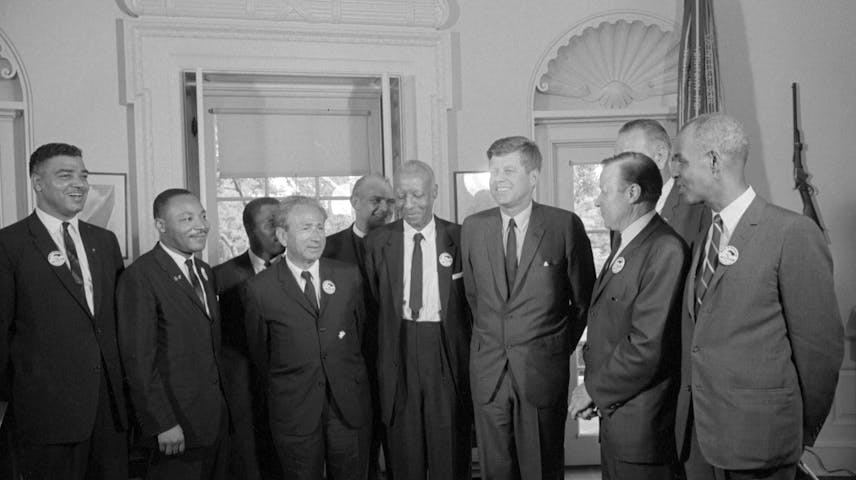 A black-and-white photograph from the 1960s showing a group of men in suits gathered together indoors, smiling and talking. Several of the men are wearing small round buttons or badges on their lapels. It is in the white house with tall windows and decorative door frames.