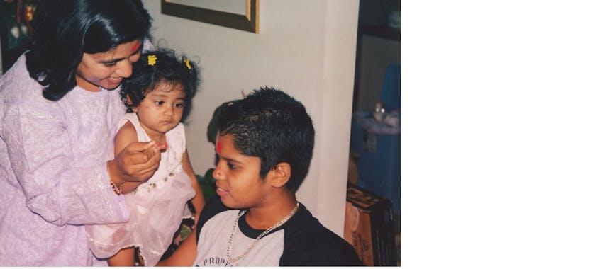 A smiling Indian American mother holding a baby up in front of an older boy as part of a Raksha Bandhan ceremony.