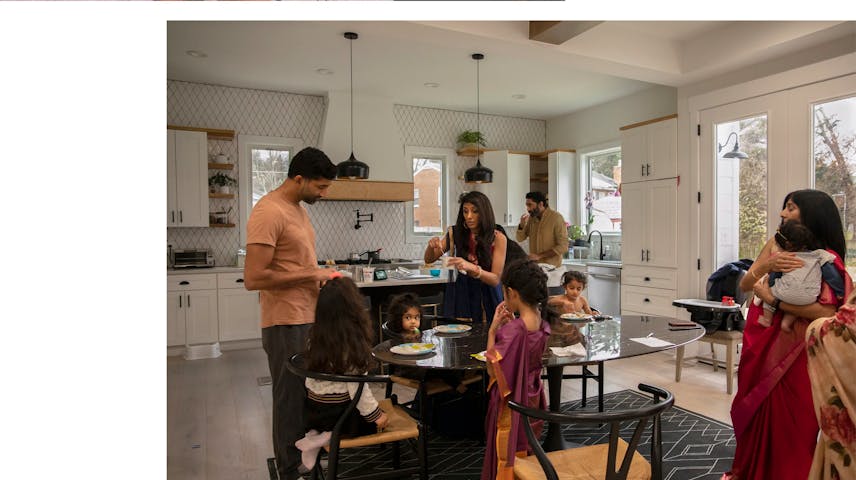 Multiple generations of an Indian American family preparing to sit down for a meal in their kitchen.