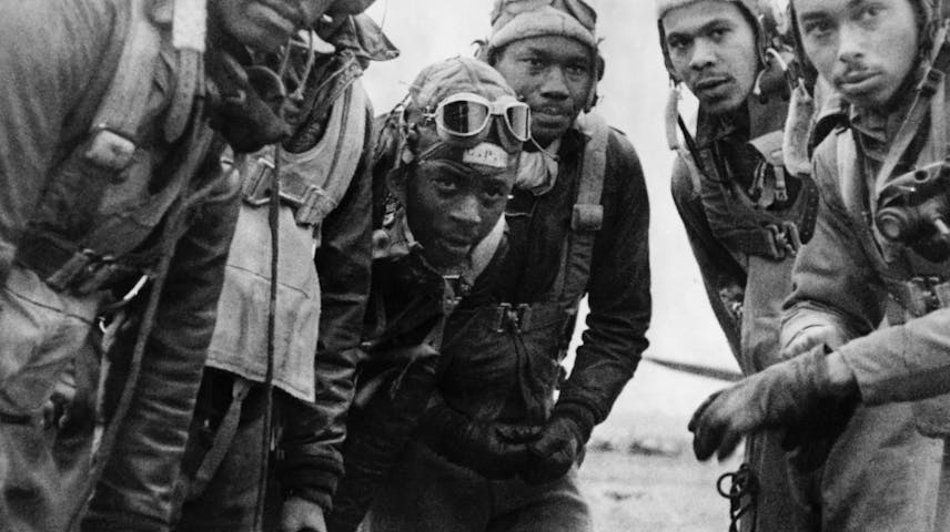 A black-and-white historical photograph of five young Black men in World War II-era flight gear, including leather helmets, goggles, flight suits, and harnesses. They are huddled closely together and looking at the camera. They were part of the Tuskegee Airmen — the first Black military pilots to serve in the U.S. Armed Forces during World War II.