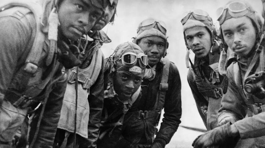 A black-and-white historical photograph of five young Black men in World War II-era flight gear, including leather helmets, goggles, flight suits, and harnesses. They are huddled closely together and looking at the camera. They were part of the Tuskegee Airmen — the first Black military pilots to serve in the U.S. Armed Forces during World War II.