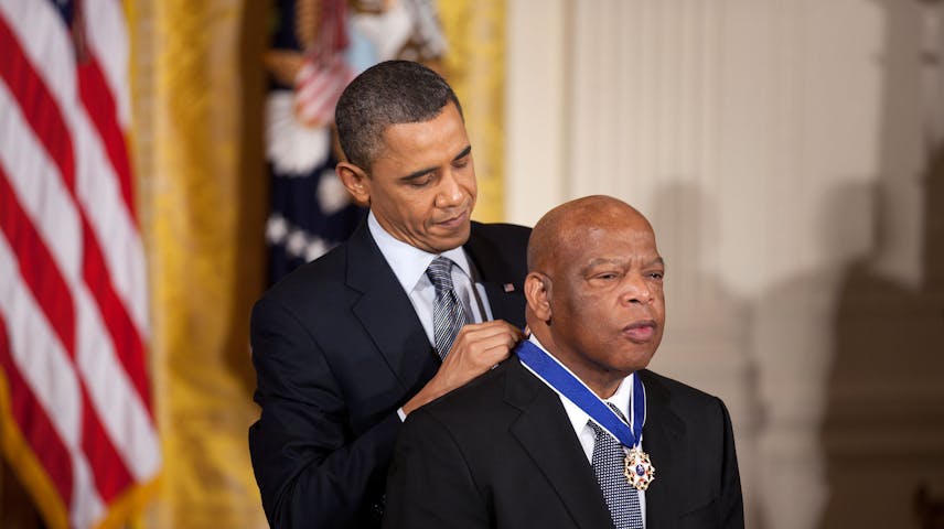A man in a dark suit places a medal on a blue ribbon around the neck of an older man in a dark suit, during a formal ceremony inside a room decorated with an American flag and gold curtains.