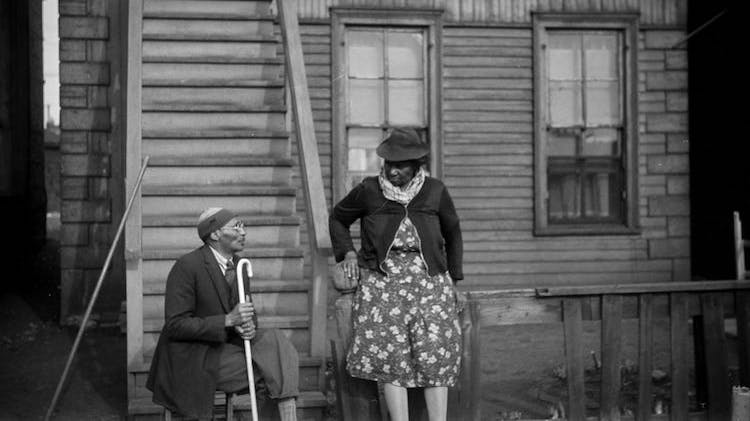 A black and white photo taken in 1941 showing a Black man and woman outside of a two-story wooden house with two windows on each floor and a set of wooden stairs that goes directly to the second floor. The woman leans against a fence that is missing some pickets, and the man sits on a stool looking up at her. He wears glasses and holds a cane, and she wears a floral dress and dark cardigan sweater.