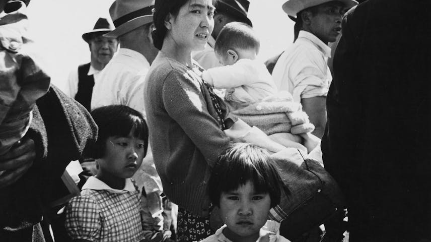 A black-and-white photograph showing a Japanese American woman holding a baby, surrounded by two young girls and a crowd of people. The woman looks stressed or worried. Everyone is dressed in 1940s clothing.