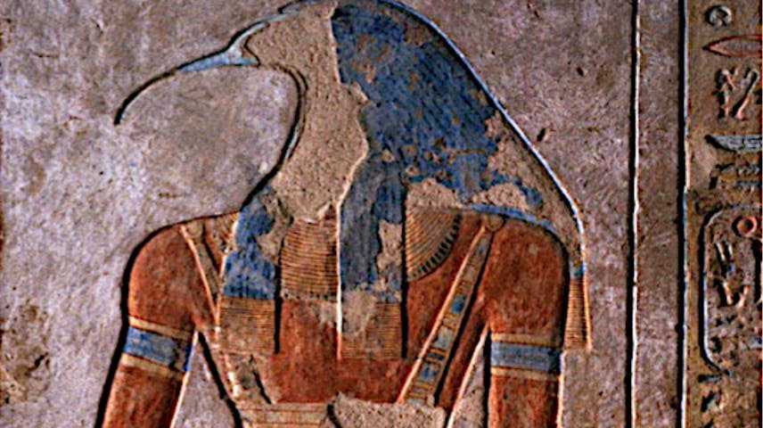 A colorful ancient Egyptian wall carving showing Thoth, the god of wisdom and writing. He has the body of a man with reddish-brown skin and wears decorated jewelry and a kilt, but his head is that of an ibis—a bird with a long, curved beak. On top of his head sits a large circular moon disk with a crescent moon beneath it, which are his symbols. Hieroglyphics (ancient Egyptian picture-writing) are carved along the right side of the image. The painting still shows its original blues, reds and golds despite being thousands of years old.