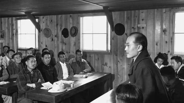 A black-and-white photograph showing a man standing at the front of a wooden room, speaking to a group of people seated at long tables who are listening attentively. The room has plain wood-paneled walls and simple windows. The photo appears to be from the mid-20th century.