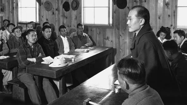 A black-and-white photograph showing a man standing at the front of a wooden room, speaking to a group of people seated at long tables who are listening attentively. The room has plain wood-paneled walls and simple windows.