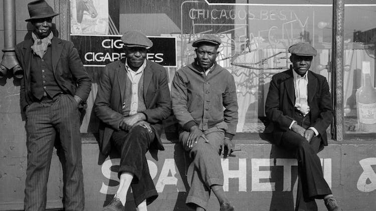 A black and white photo taken in 1941 showing four middle-aged Black men facing into the sun while three are perched on the outside windowsill of a restaurant and the fourth leans on the wall next to them. The word “spaghetti” is painted on the wall underneath the window and is visible through their legs.