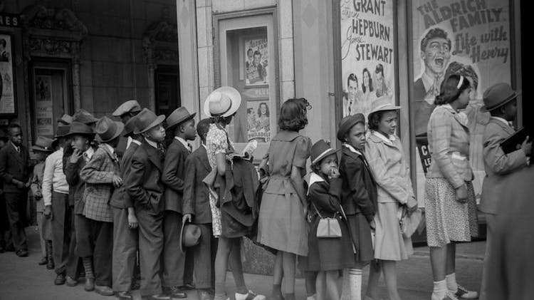 A black and white photo taken in 1941 showing nearly 20 Black children in line for a movie theatre. The boys wear suits, hats and dress shoes, and the girls wear suits or dresses, saddle shoes or heels, and hats.