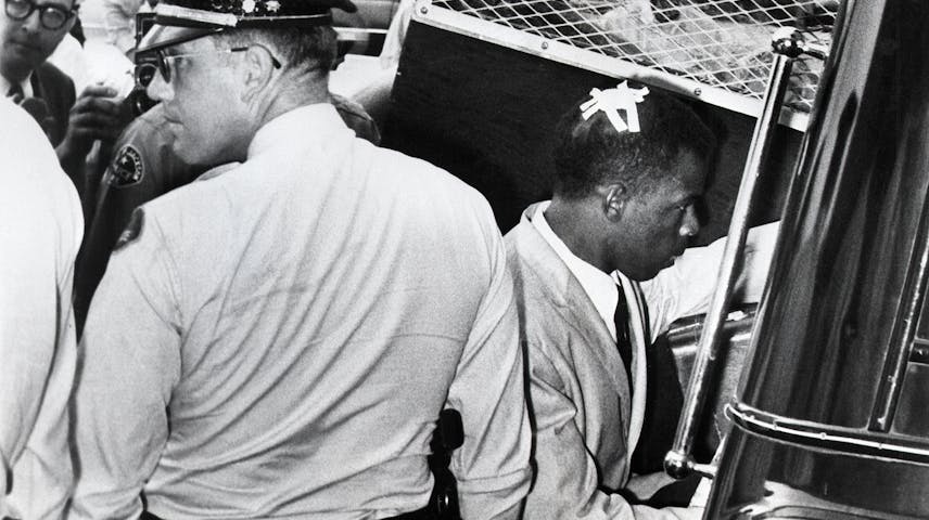 A black-and-white historical photograph showing a young Black man in a dress shirt and tie being placed into a police vehicle by a white police officer. The man has a bandage on his head. Other people and a photographer can be seen in the background. The image appears to be from the Civil Rights era of the 1950s or 1960s.