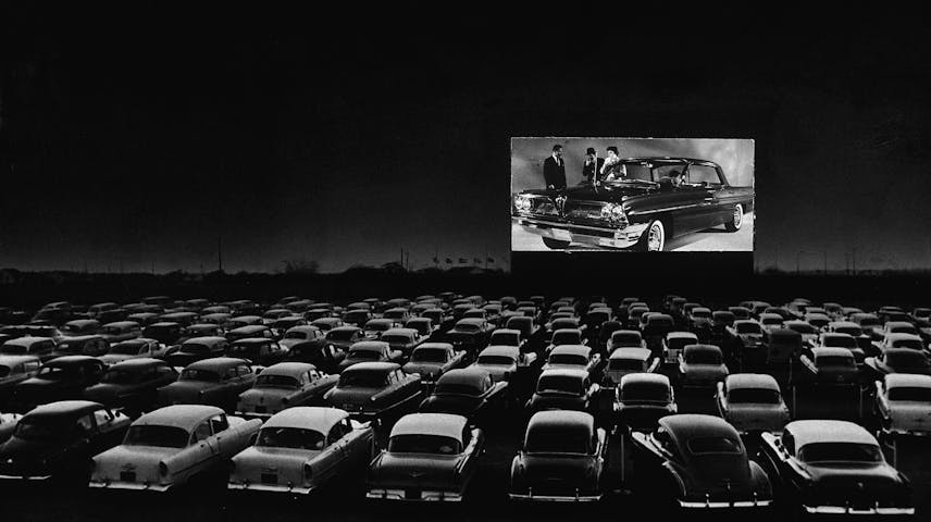 A black and white photo of a packed drive-in movie theater at night. Dozens of classic 1950s cars fill a large outdoor lot, all facing a big screen in the distance that shows a scene with people standing next to a fancy car. The sky is dark, and the glowing screen is the brightest thing in the image.