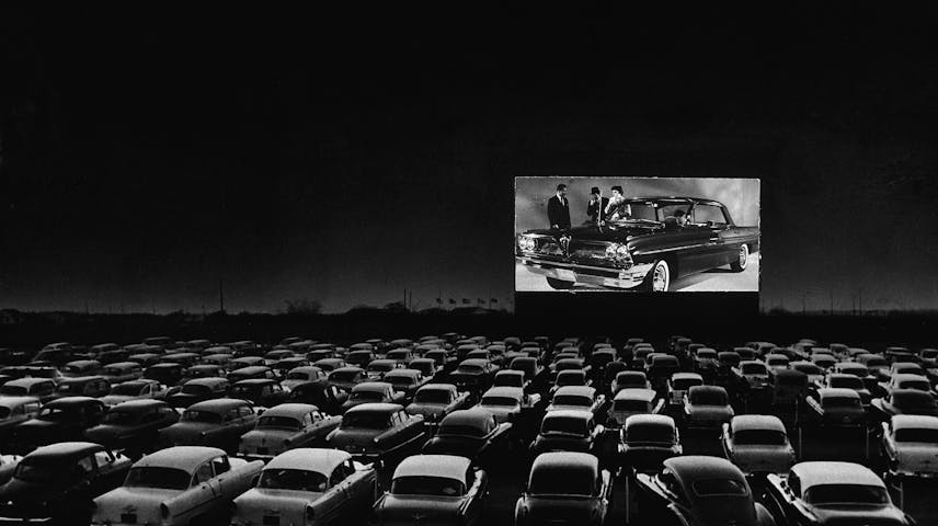 A black and white photo of a packed drive-in movie theater at night. Dozens of classic 1950s cars fill a large outdoor lot, all facing a big screen in the distance that shows a scene with people standing next to a fancy car. The sky is dark, and the glowing screen is the brightest thing in the image.
