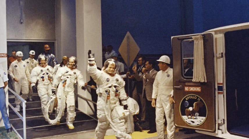 Three astronauts (Neil A. Armstrong, Michael Collins and Edwin E. Aldrin Jr.) leave a building to enter an awaiting vehicle whose door is labeled “Apollo 11.” The astronaut in front waves to onlookers.