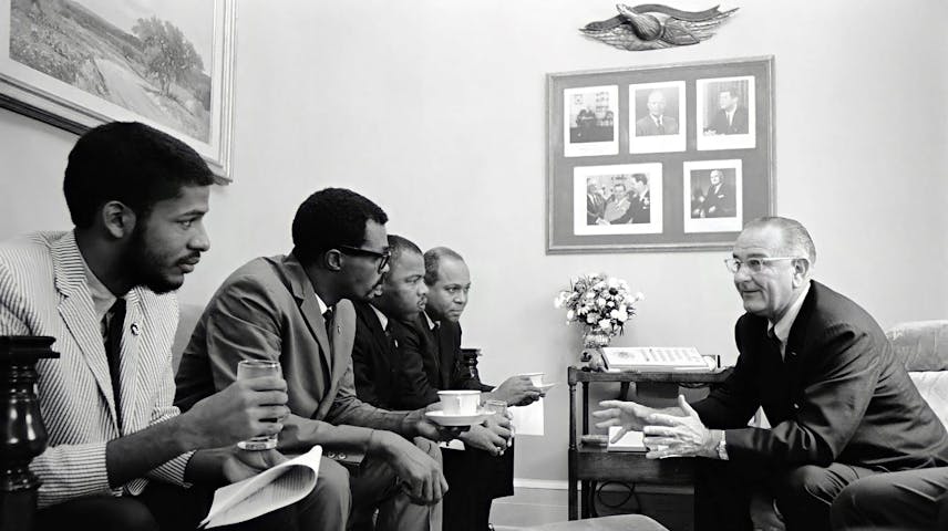 A black-and-white photo showing four young Black men seated in a row, dressed in suits and casual clothing, meeting with an older white man in a suit who is speaking and gesturing with his hands. President Johnson is the white man and the civil rights activists John Lewis and James Farmer are seated third and fourth to the right. They are seated in what appears to be a formal office or meeting room. A framed collage of portraits hangs on the wall in the background, and a small table with flowers sits between the two groups.