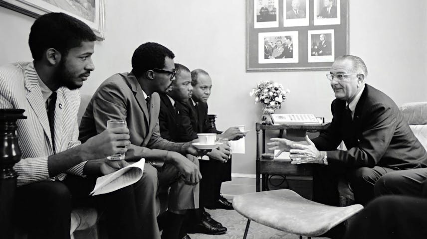 A black-and-white photo showing four young Black men seated in a row, dressed in suits and casual clothing, meeting with an older white man in a suit who is speaking and gesturing with his hands. President Johnson is the white man and the civil rights activists John Lewis and James Farmer are seated third and fourth to the right. They are seated in what appears to be a formal office or meeting room. A framed collage of portraits hangs on the wall in the background, and a small table with flowers sits between the two groups.