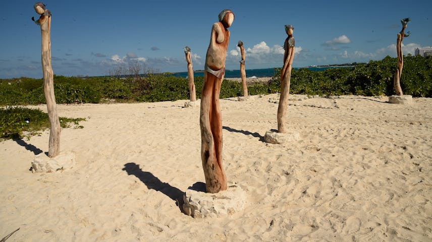 Several tall wooden sculptures carved to look like slender human figures stand in the white sand of a tropical beach. The figures are made from natural driftwood and have simple, abstract bodies with small round heads. Green bushes and bright blue ocean water are visible in the background under a sunny sky.