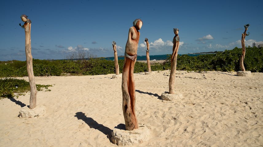 Several tall wooden sculptures carved to look like slender human figures stand in the white sand of a tropical beach. The figures are made from natural driftwood and have simple, abstract bodies with small round heads. Green bushes and bright blue ocean water are visible in the background under a sunny sky.
