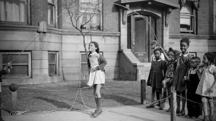 A black and white photo taken in 1941 showing Black children playing jump rope together on the sidewalk in front of a brick building. One girl is jumping, two children are swinging the rope, and several others watch. Two of the girls are smiling at the camera.