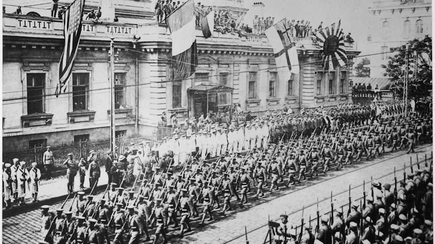 A black and white historical photograph showing hundreds of soldiers in uniform and armed with rifles marching in formation down a wide city street. Several flags are displayed on a large government-style building in the background, including the American flag, the French flag, a British naval flag and the Japanese Imperial rising sun flag. Crowds of spectators and additional military personnel watch from the sidewalks and rooftops.