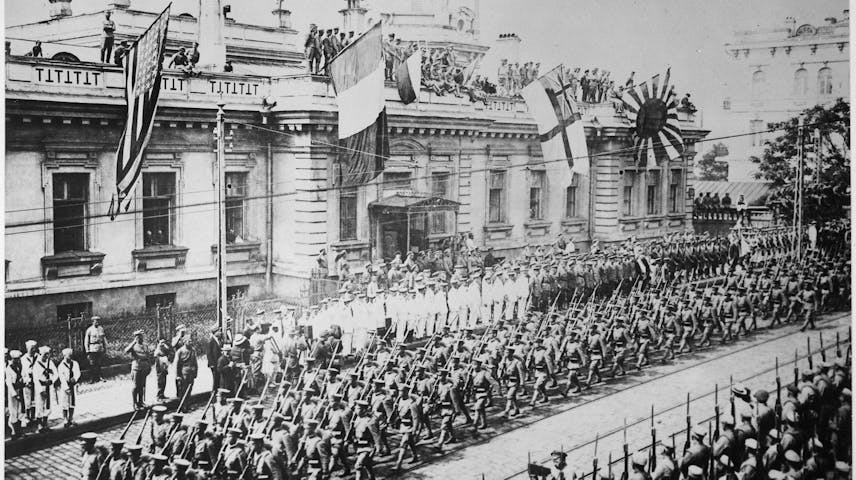 A black and white historical photograph showing hundreds of soldiers in uniform and armed with rifles marching in formation down a wide city street. Several flags are displayed on a large government-style building in the background, including the American flag, the French flag, a British naval flag and the Japanese Imperial rising sun flag. Crowds of spectators and additional military personnel watch from the sidewalks and rooftops.