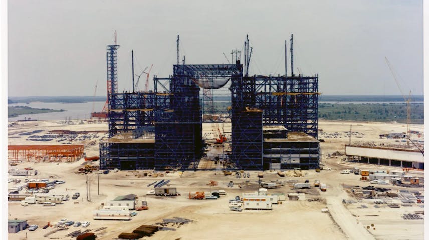 Photograph of a large structure under construction. In front of it and to the sides is flat land with building supplies, vehicles and trailers, and behind it is open water and marshland.