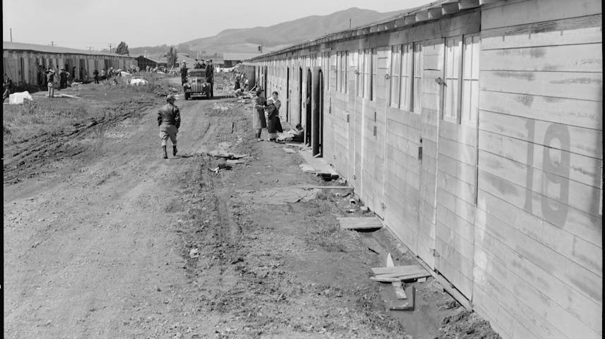 A black-and-white photograph showing a row of plain wooden barracks buildings in California during World War II. A muddy dirt road runs alongside the buildings. Several people, including a soldier and civilians, stand near the barracks doors. Mountains are visible in the background. The conditions look crowded and basic, reflecting the difficult living situation faced by Japanese Americans who were forced to live here after the U.S. government ordered their removal from their homes following the attack on Pearl Harbor in 1941