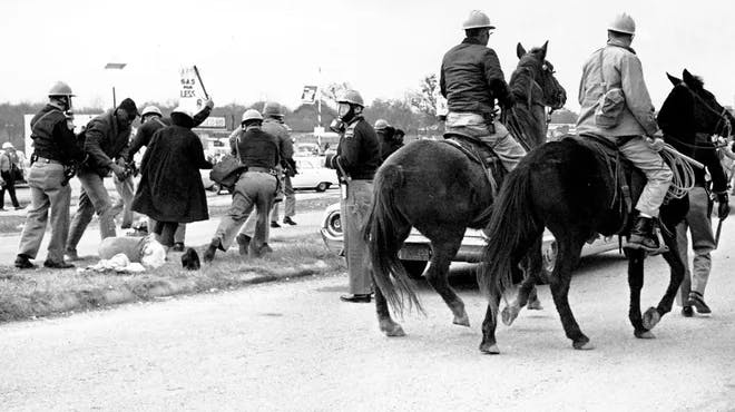 A black-and-white historical photograph showing police officers on horseback and on foot using force against civil rights protesters on a road. Some protesters can be seen on the ground. One protester is holding a sign.