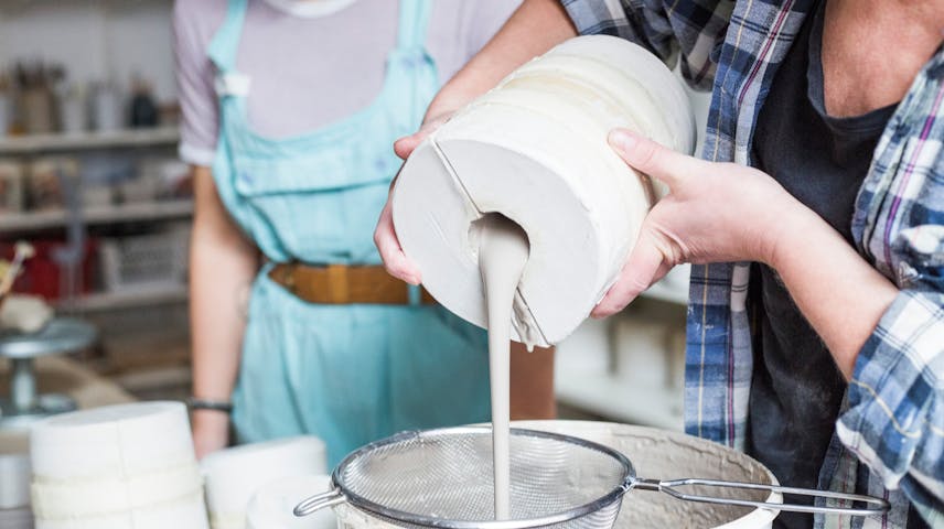 A potter pours liquid clay from a large vase through a sieve resting on top of a bucket.