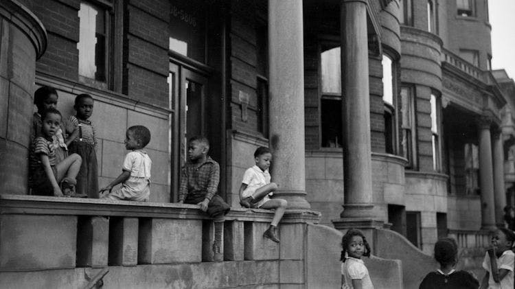 A black and white photo taken in 1941 showing several Black children sitting and standing on and around the front stoop of a brick building on a city street